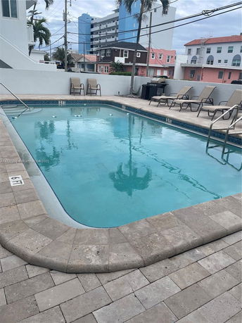 Outdoor swimming pool surrounded by lounge chairs and a small wall, with buildings in the background.
