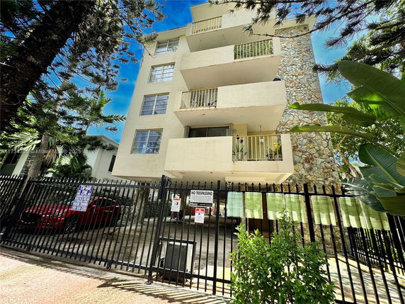 Front view of a multi-story building with balconies and a stone facade, behind a black metal fence.