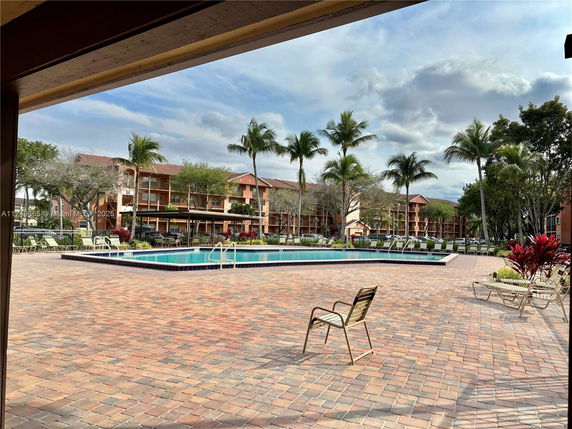 Outdoor swimming pool area with chairs and surrounded by palm trees and multi-story buildings.