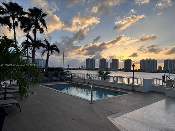 Outdoor swimming pool area with lounge chairs and a view of high-rise buildings across the water at sunset.