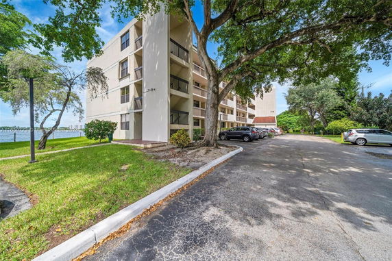 Parking area beside a multi-story residential building with surrounding trees and partial lake view.