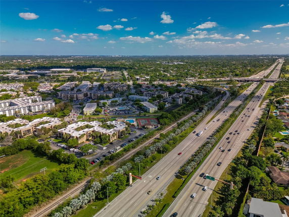 Aerial view of a highway with multiple lanes, surrounded by residential and commercial buildings, green areas, and a distant horizon under a blue sky.