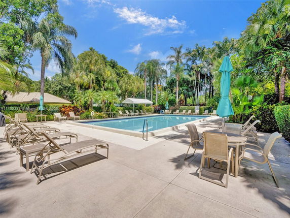 Outdoor area featuring a swimming pool surrounded by chairs and tables with umbrellas, set amidst lush greenery.