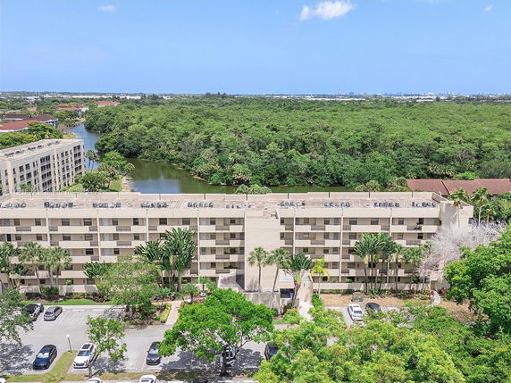 Aerial view of a multi-story building with a rooftop featuring air conditioning units, surrounded by greenery and water in the background.