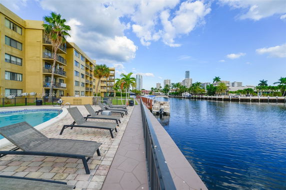 Outdoor swimming pool area with lounge chairs alongside a waterfront and multi-story building.