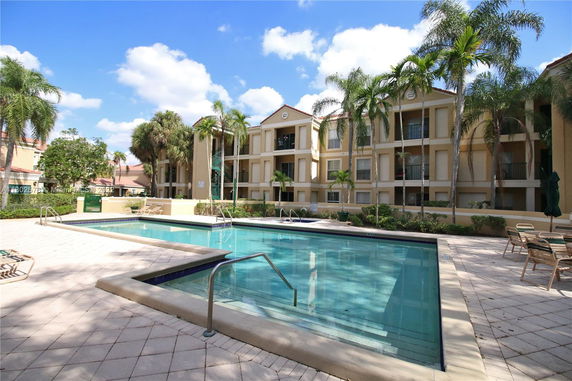 Outdoor swimming pool area in front of a multi-story residential building with palm trees.