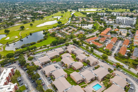 Aerial view of a residential area with multiple rows of houses, a swimming pool, parking areas, and a golf course with lakes in the background.