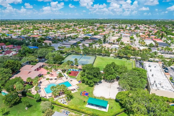 Panoramic view of a residential neighborhood with a pool, tennis courts, greenery, and surrounding houses.