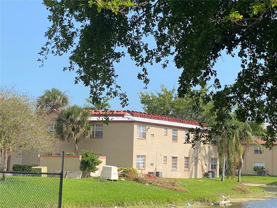 Front view of a two-story apartment building with a red tile roof and beige exterior walls, surrounded by trees and grass.