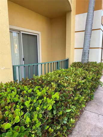 Exterior view of a building with light yellow walls, a sliding glass door, a railing in front of a small patio area, and surrounding green bushes.