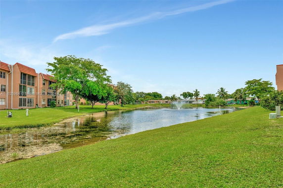 View of apartment buildings next to a water body with fountain and green lawns.