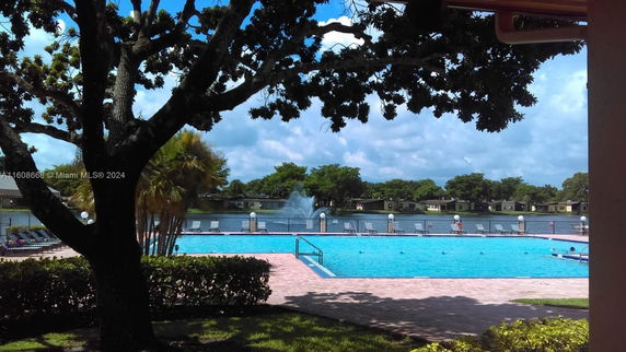 View of an outdoor swimming pool with lounge chairs next to a lake and a fountain.