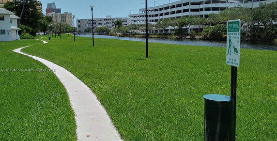 Outdoor view with a sidewalk, grassy area, and a river alongside buildings in the background.