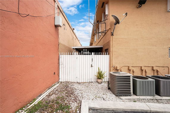 Exterior view of a narrow alleyway between two buildings with air conditioning units and a white gate.