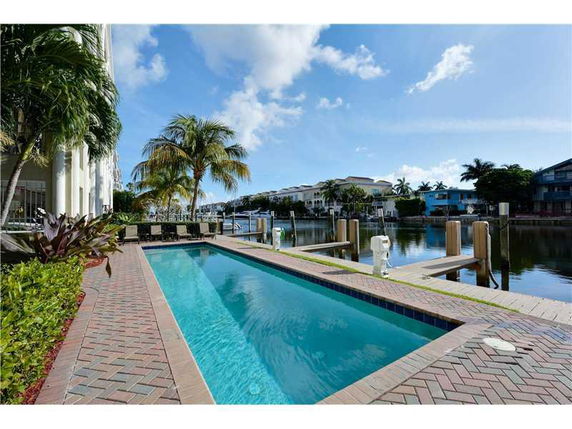 View of a narrow swimming pool next to a canal with houses and palm trees in the background.