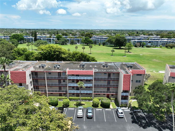 Aerial view of a four-story residential building with parking area in front and green open spaces behind.