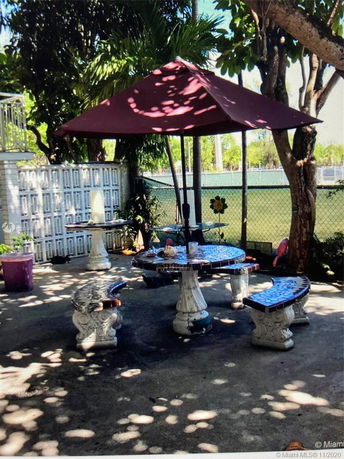 Outdoor area with a round table, benches, and a red umbrella near a fenced tennis court.