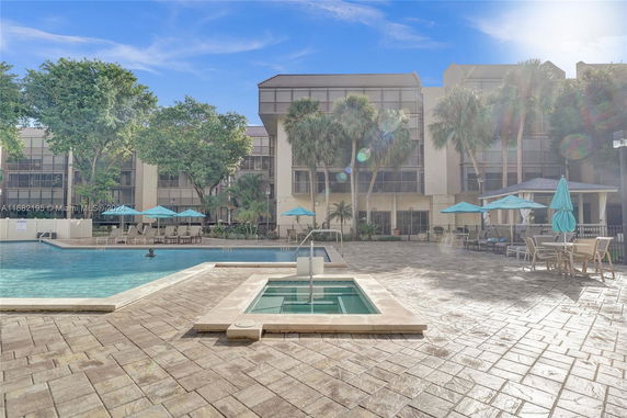 Outdoor swimming pool area with lounge chairs and umbrellas in front of a multi-story building.