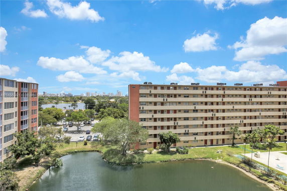A multi-story residential building seen from a distance, with a parking area and a pond in front.
