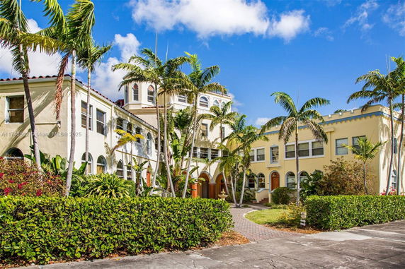 Front view of a multi-story building with arched windows and a tower, surrounded by palm trees.