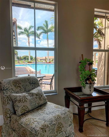 View of a swimming pool area through a window, with palm trees and outdoor seating visible.