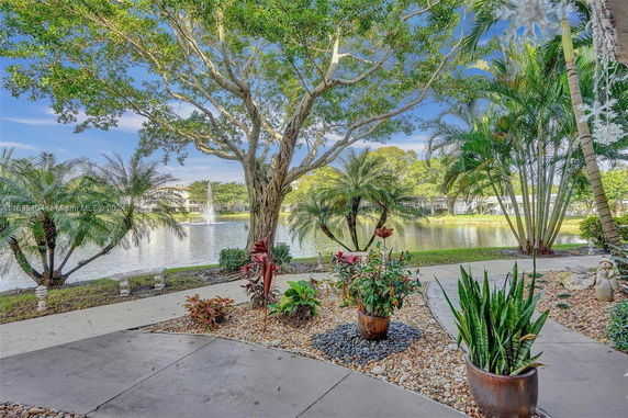 View of a landscaped area with potted plants and trees by a lake with a fountain.