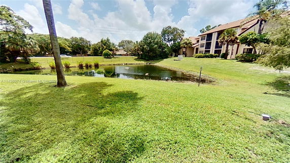 View of a pond surrounded by grassy areas with trees and a building in the background.