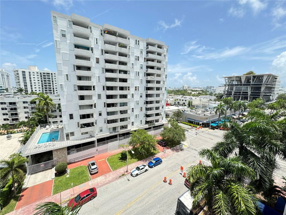 View of a multi-story apartment building with a swimming pool and surrounding area including streets and other buildings.