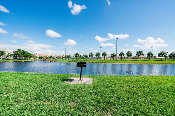 View of a grassy area with a small lake and buildings in the background under a blue sky.