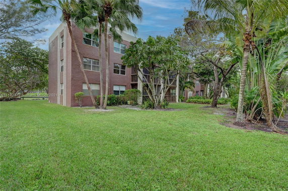 Front view of a multi-story apartment building with a brown exterior and large windows, surrounded by greenery and trees.