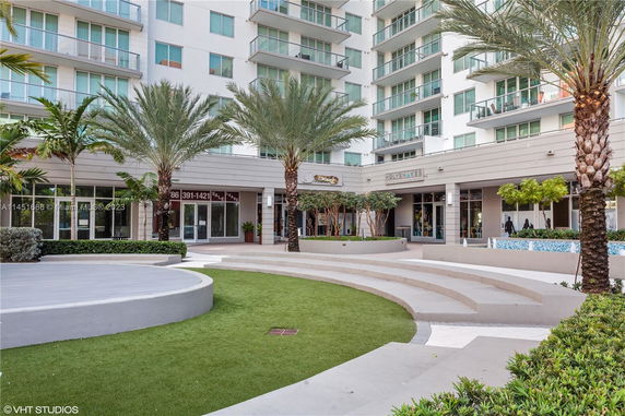 Front view of a modern multi-story building with shops on the ground floor and a landscaped outdoor area.