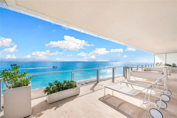Wide angle view from a balcony overlooking the ocean with lounge chairs and plants.