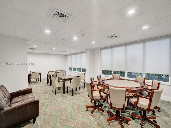 Room with tables and chairs, carpeted floor, and large windows with blinds.