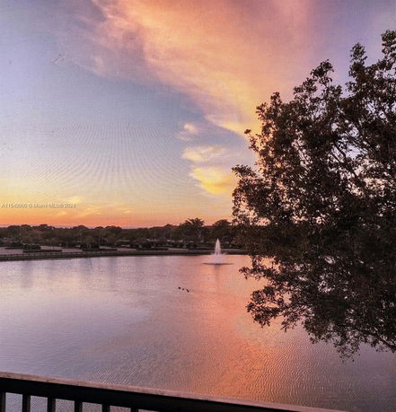 View of a lake with a fountain and trees, under a colorful sky at sunset.