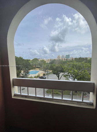 View through a balcony archway of surrounding foliage and distant buildings with a swimming pool visible below.