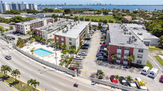 Aerial view of apartment buildings with parking area and swimming pool, surrounded by roads and trees, with a distant view of the city and water.