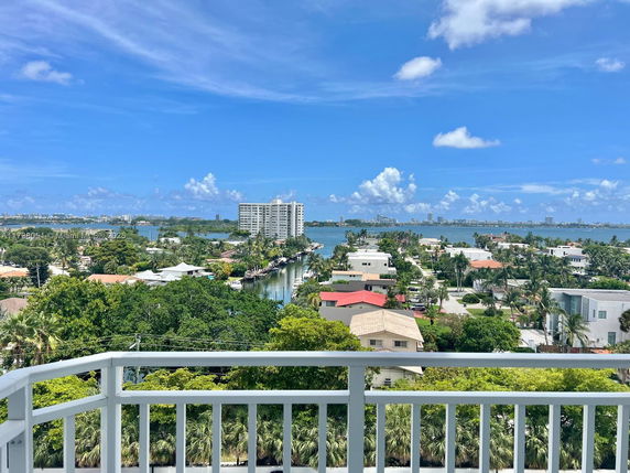 Panoramic view from a balcony overlooking houses, greenery, and a body of water under a blue sky.