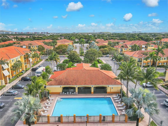 Aerial view of a residential complex with a large swimming pool at the center, surrounded by palm trees and buildings with red-tiled roofs.