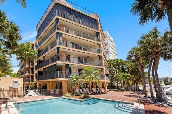 Exterior view of a multi-story building with balconies, adjacent to a swimming pool area surrounded by palm trees.