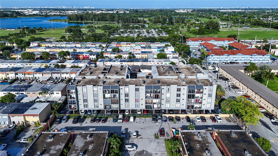Aerial view of a residential complex with multiple buildings and parking areas, surrounded by greenery and other residential units.