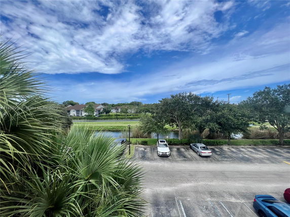 View of a parking area with cars and distant buildings surrounded by trees and greenery.