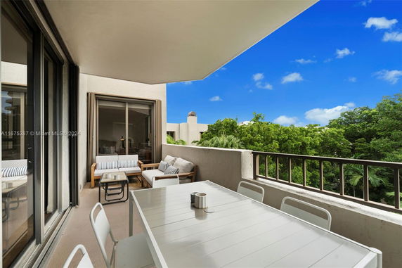 Balcony with outdoor seating and table, overlooking green trees under a blue sky.