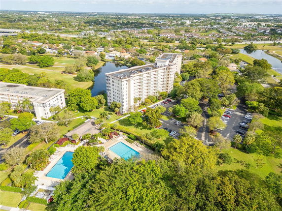 Aerial view of a multi-story residential building with adjacent swimming pools and parking areas, surrounded by greenery and other residential areas in the distance.