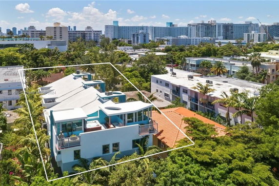 Aerial view of a building with a rooftop featuring an open terrace and a blue exterior, surrounded by trees and other buildings.