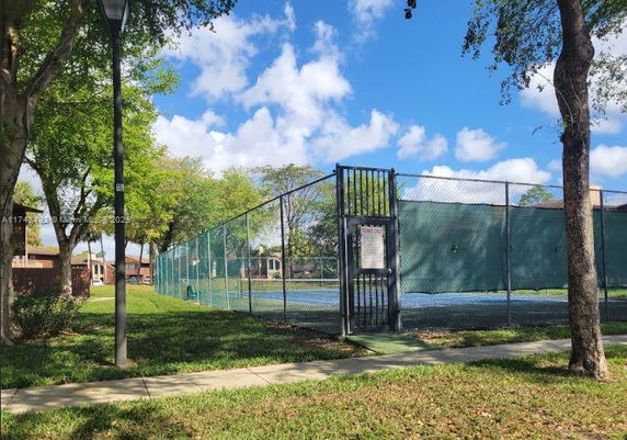 View of a tennis court surrounded by a fence, with trees and a pathway nearby.