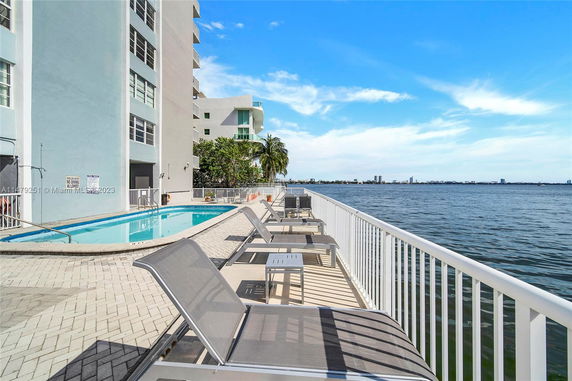 Outdoor swimming pool area adjacent to a building with lounge chairs and a view of the water.