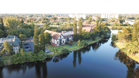 Aerial view showing a building complex near a body of water with surrounding trees and parking area.