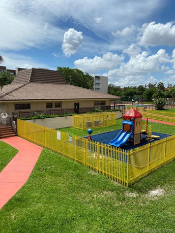 View of a playground with a play structure and fenced area beside a building with a sloped roof.
