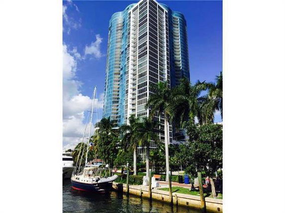 View of a tall modern building with glass facade next to a waterfront, surrounded by palm trees.