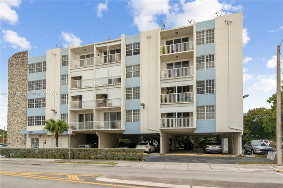 Front view of a multi-story apartment building with balconies and parking on the ground level.